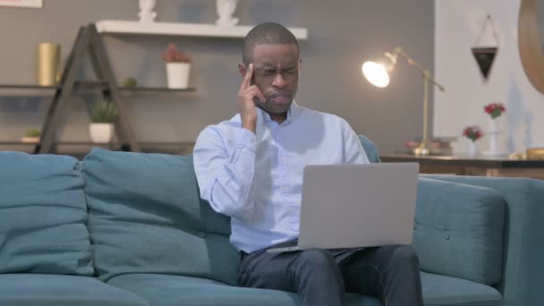 Hombre africano con portátil con dolor de cabeza en el sofá — Vídeos de Stock