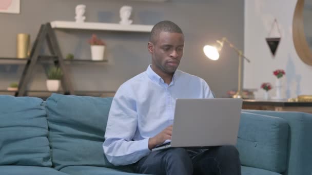 Hombre africano con portátil reaccionando a la pérdida en el sofá — Vídeos de Stock