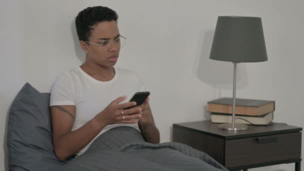 ベッドのラップトップを使用している間にスマートフォンで話す怒っているアフリカの女性 — ストック動画