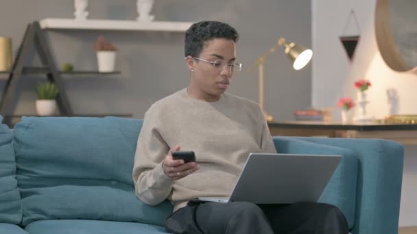 ソファの上のスマートフォンを使用してノートパソコンを持つアフリカの女性 — ストック動画
