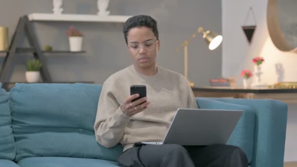 Arg afrikansk kvinna med bärbar dator Talar på smartphone på soffan — Stockvideo