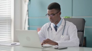 Ofiste dizüstü bilgisayar kullanırken kameraya gülümseyen genç Afrikalı Doktor