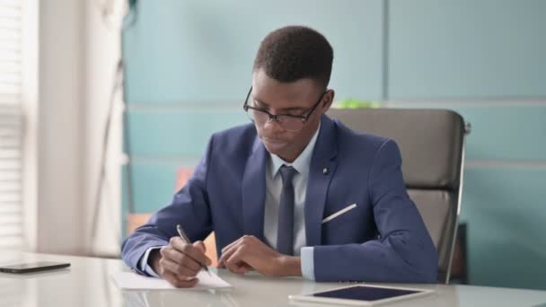 Молодой африканский бизнесмен пишет на бумаге в офисе — стоковое видео