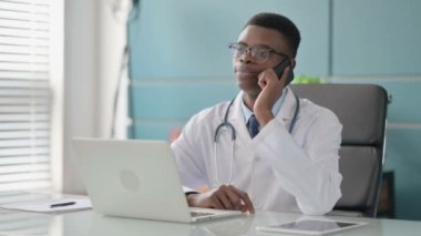Ofiste dizüstü bilgisayar kullanırken telefonda konuşan genç Afrikalı Doktor