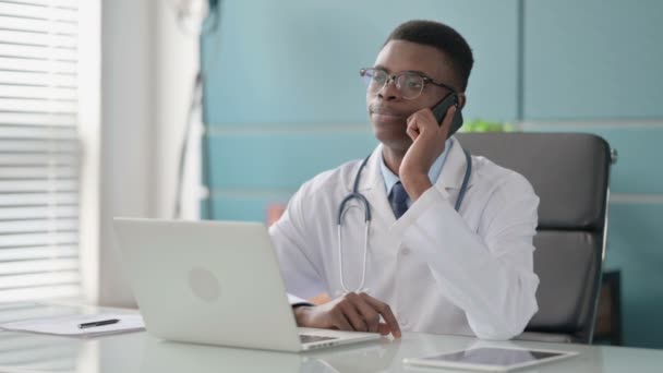 Молодой африканский врач разговаривает по телефону во время использования ноутбука в офисе — стоковое видео