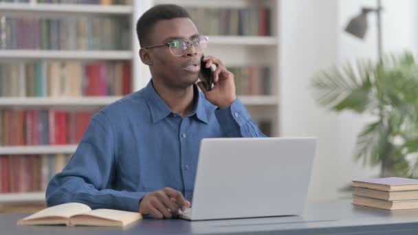 Jeune homme africain parlant au téléphone tout en utilisant un ordinateur portable dans le bureau — Video