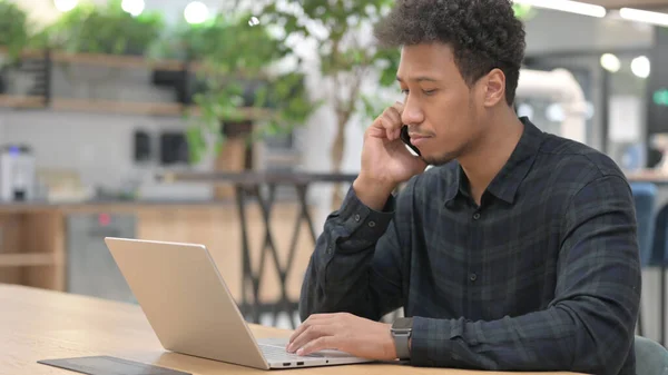 一个拿着笔记本电脑用智能手机说话的非洲裔美国人 — 图库照片