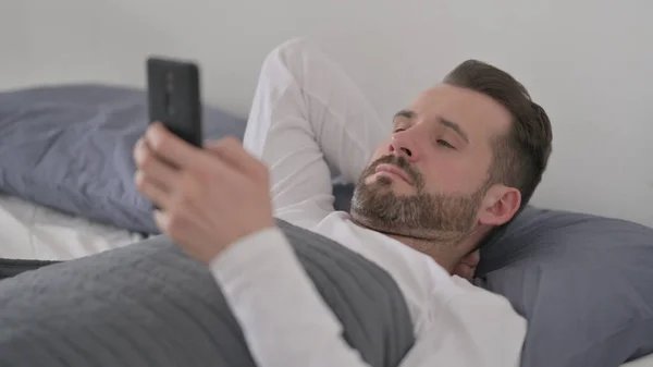 Homem usando Smartphone enquanto deitado na cama — Fotografia de Stock