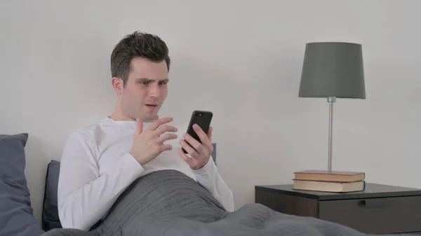 Homem reagindo a perda no smartphone na cama — Fotografia de Stock