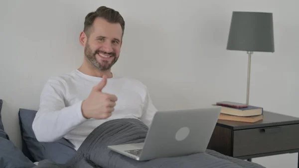 Dizüstü bilgisayarlı adam Başparmakları Yatakta Gösteriyor — Stok fotoğraf