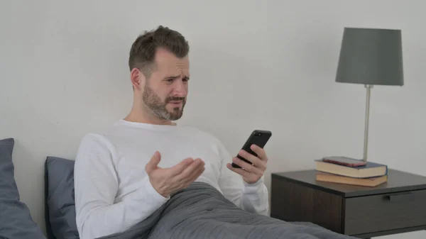 Mężczyzna reaguje na utratę na smartfonie w łóżku — Zdjęcie stockowe