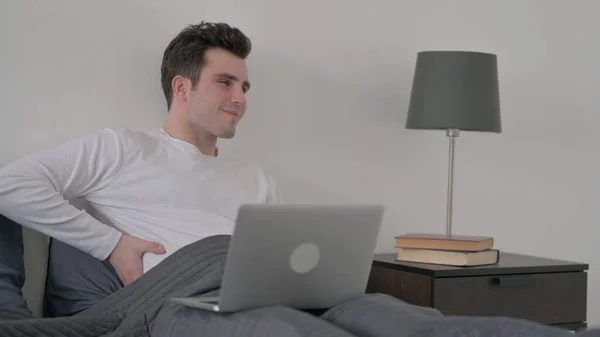 Hombre con dolor de espalda trabajando en el ordenador portátil en la cama —  Fotos de Stock