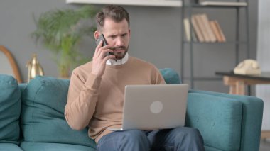 Dizüstü bilgisayarlı kızgın adam telefonda konuşuyor. 