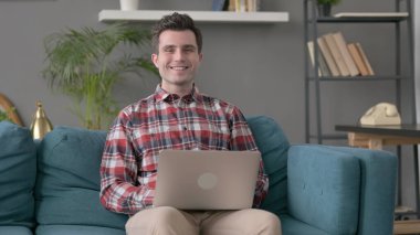 Dizüstü bilgisayarlı bir adam kanepedeki kameraya gülümsüyor. 