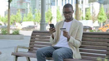 Bench 'te otururken akıllı telefondan alışveriş yapan Afrikalı Amerikalı adam.