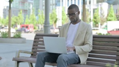 Dizüstü bilgisayarlı Afrikalı Amerikalı Adam Bench 'te otururken kameraya bakıyor.