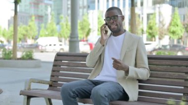 Bankta otururken telefonda konuşan Afrikalı Amerikalı adam.