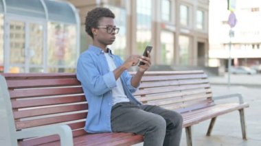 African Man Celebrating Online Success on Smartphone while Sitting Outdoor on Bench