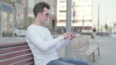 Side View of Casual Man Using Smartphone while Sitting Outdoor on Bench