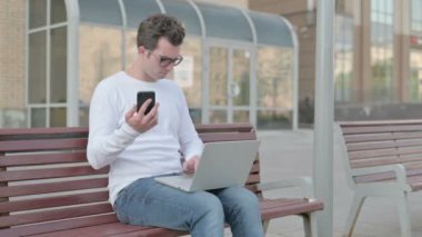 Casual Man Using Smartphone and Laptop while Sitting Outdoor on Bench