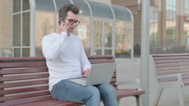 Angry Casual Man Talking on Phone and using Laptop while Sitting Outdoor on Bench