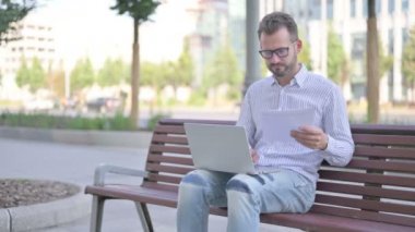 Adult Man Reading Documents and Working on Laptop while Sitting on Bench Outdoor