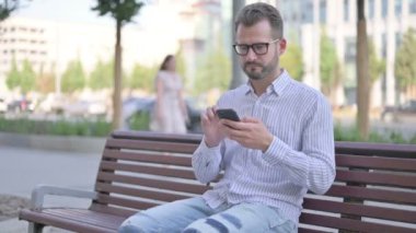 Adult Man Celebrating Online Success on Smartphone while Sitting Outdoor on Bench