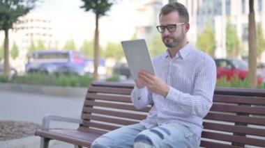 Adult Man Celebrating Online Win on Tablet while Sitting Outdoor on Bench