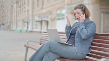 Angry Senior Old Woman Talking on Phone and using Laptop while Sitting Outdoor on Bench