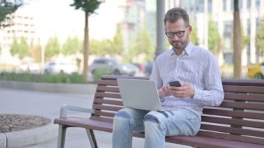 Adult Man Talking on Phone and using Laptop while Sitting Outdoor on Bench