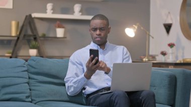 The Young African Man using Smartphone while using Laptop on Sofa