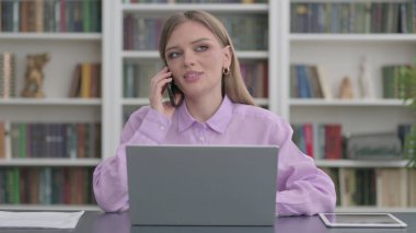 Ofiste dizüstü bilgisayar kullanan genç bir kadın telefonda konuşuyor.
