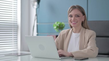 Ofiste dizüstü bilgisayar kullanırken kameraya gülümseyen genç iş kadını