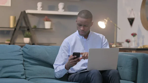 Young African Man Using Smartphone While Using Laptop Sofa — 스톡 사진