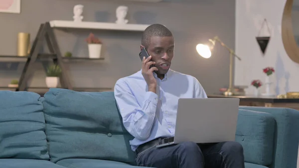 Young African Man Talking Smartphone While Using Laptop Sofa —  Fotos de Stock