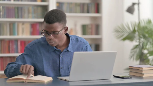 African Man Reading Book and Working on Laptop in Office