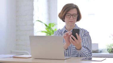 Yaşlı kadın dizüstü bilgisayar kullanırken akıllı telefon kullanıyor