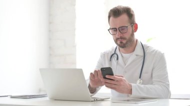 Middle Aged Doctor using Smartphone while using Laptop in Clinic