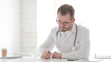 Middle Aged Doctor Thinking while Writing on Paper in Clinic