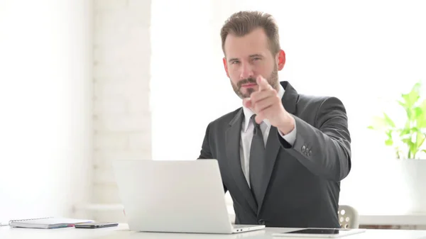 Businessman Pointing at Camera while using Laptop in Office