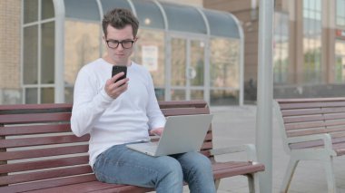 Casual Man Using Smartphone and Laptop while Sitting Outdoor on Bench