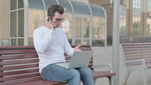 Casual Man Talking on Phone and using Laptop while Sitting Outdoor on Bench