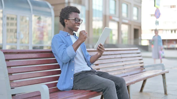 African Man Celebrating Online Win on Tablet while Sitting Outdoor on Bench