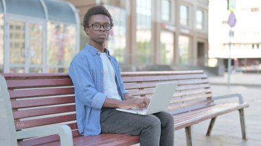 Dizüstü bilgisayarlı Afrikalı Adam Bench 'te otururken kameraya bakıyor.