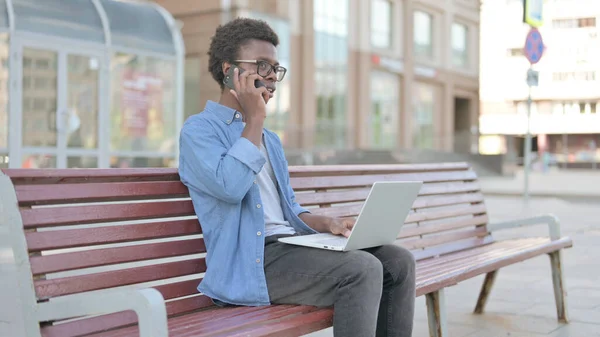 African Man Talking on Phone and using Laptop while Sitting Outdoor on Bench