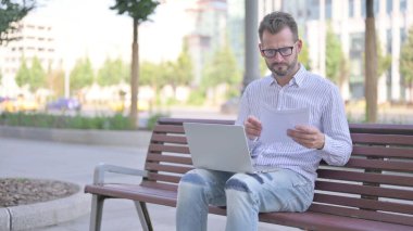 Adult Man Reading Documents and Working on Laptop while Sitting on Bench Outdoor
