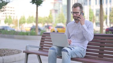 Adult Man Talking on Phone and using Laptop while Sitting Outdoor on Bench
