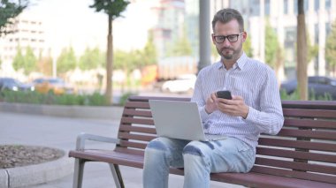 Adult Man Using Smartphone and Laptop while Sitting Outdoor on Bench