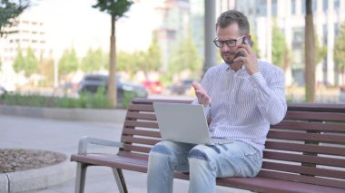 Angry Adult Man Talking on Phone and using Laptop while Sitting Outdoor on Bench