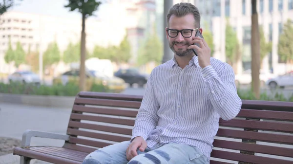 Adult Man Talking on Phone while Sitting Outdoor on Bench